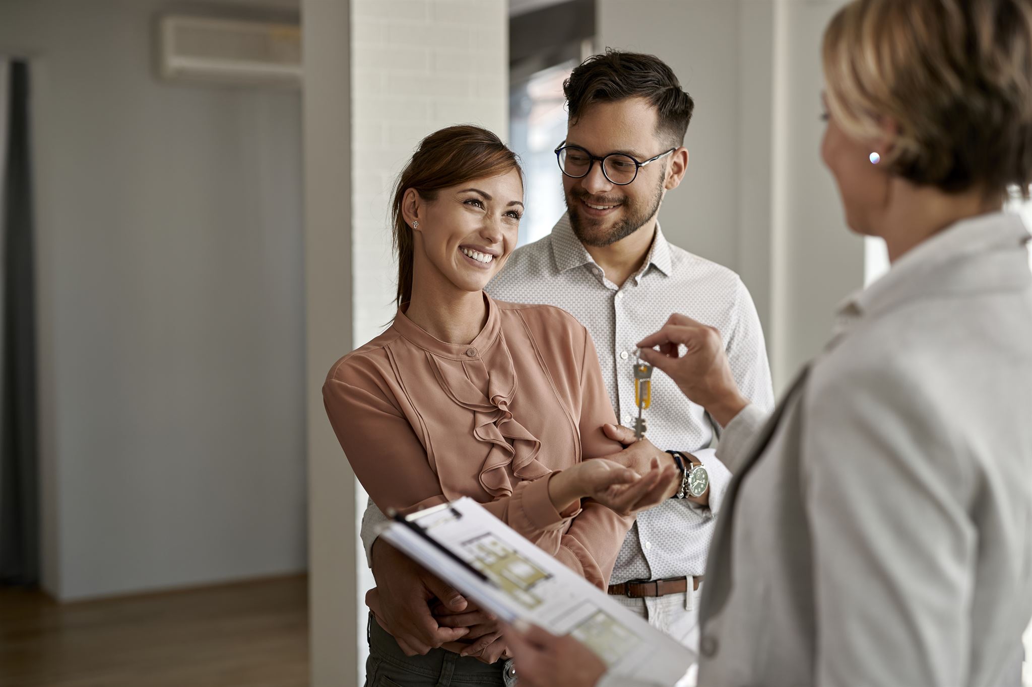 Woman-in-business-suit,-holding-clipboard-with-floorplan,-handing-key-to-smiling-couple